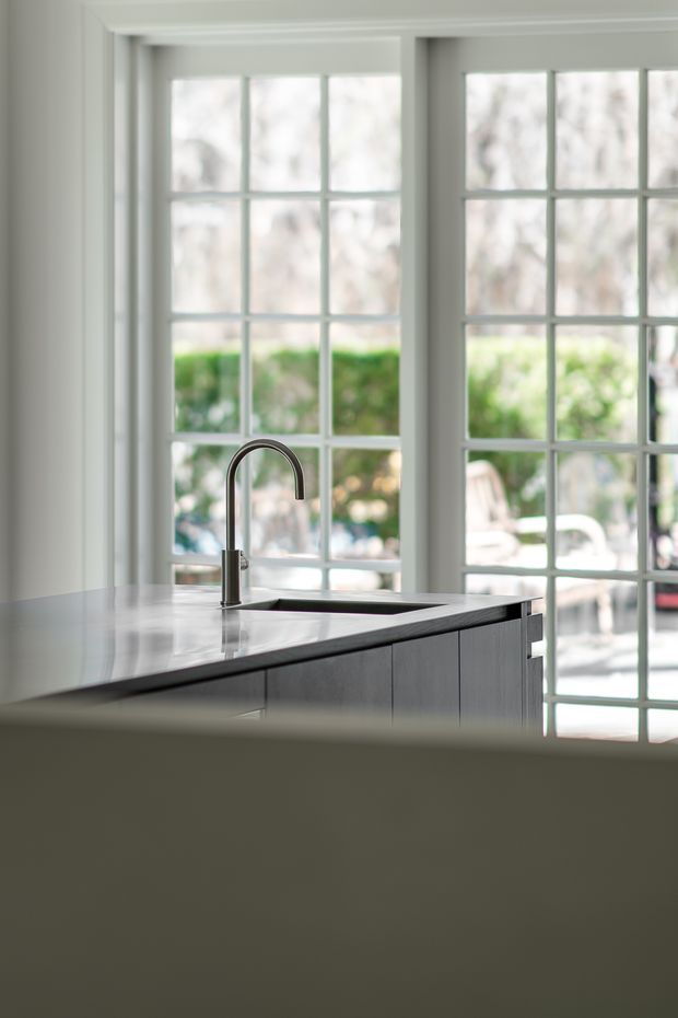 Bright, refined kitchen interior featuring a large island with a pale stone benchtop, integrated sink and curved metal tap, pale grey fixed cabinetry, and expansive white-framed multi-pane glazing overlooking greenery and an outdoor seating area.