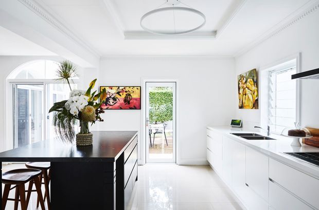Bright, refined white kitchen with a large dark island, timber stools, white cabinetry, integrated appliances, a white tiled or glossy floor, and abundant glazing. Decorative artwork and a floral arrangement add colour, while ornate ceiling cornices and a contemporary circular pendant combine classic architectural detailing with modern joinery.