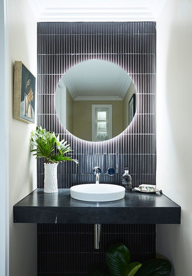 A compact, refined bathroom vanity with a round vessel basin set on a thick dark stone benchtop. The wall behind is clad in narrow glossy dark tiles arranged in vertical lines, with a large circular mirror centred above the basin. Chrome tapware, black accessories, a white sculptural vase, and green plants add contrast. A hallway and window are visible in the mirror reflection.