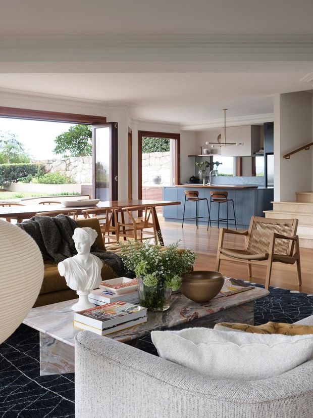 A bright, open-plan living and dining area with a visible kitchen beyond. The room features warm timber flooring, a marble coffee table, upholstered seating, a patterned dark rug, sculptural decor, and large timber-framed glazed doors opening to the garden. A blue-grey kitchen island with bar seating sits at the rear, while a staircase rises along the right wall.