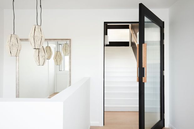 Bright minimalist interior view of a white hallway or stair landing, featuring a black-framed glazed door with a timber handle opening toward a white staircase. A large wall mirror reflects several sculptural woven pendant lights, while pale timber flooring adds warmth.