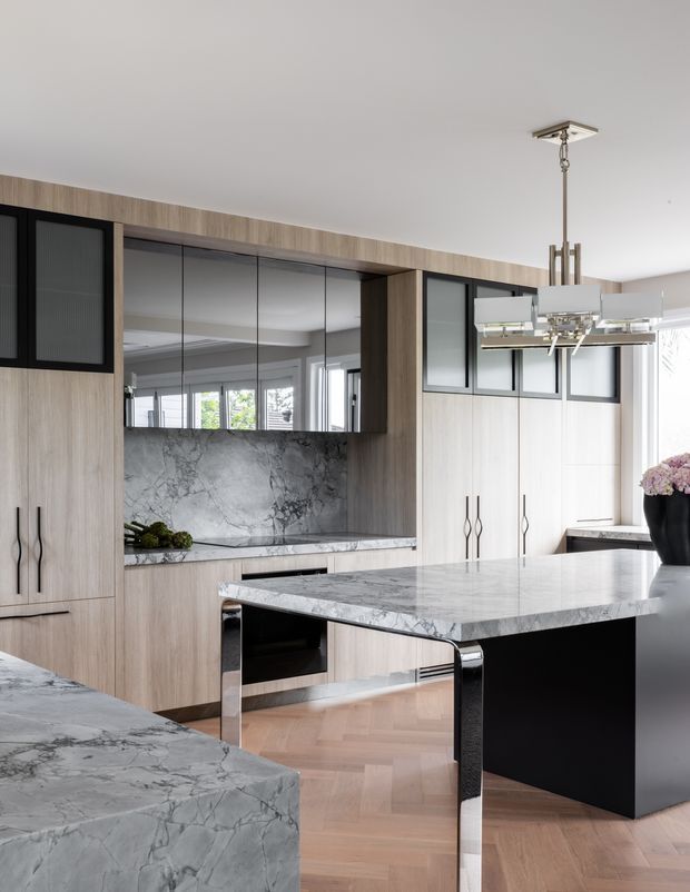 A refined contemporary kitchen with extensive light timber cabinetry, black-framed upper glass-front cabinets, mirrored backsplash panels, and dramatic white-and-grey veined stone benchtops. A large central island has a waterfall stone edge, black cabinetry, and polished chrome supports. The room includes a sculptural linear pendant light, herringbone timber flooring, and a bright windowed outlook.