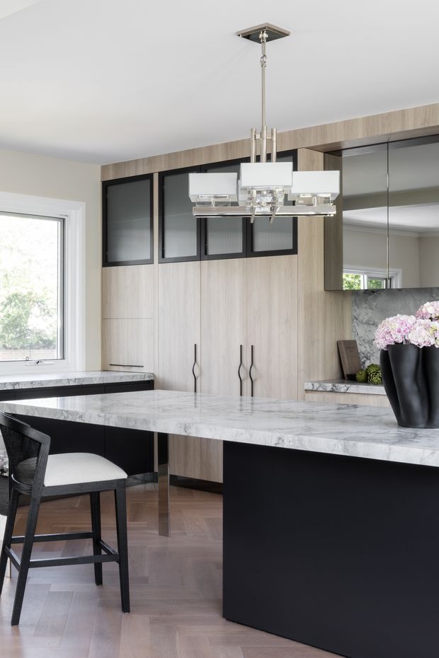 A refined contemporary kitchen featuring a large marble island with a dark matte base, light timber-look cabinetry, black-framed upper display cabinets, mirrored splashback or wall panels, and a modern geometric pendant light. A bar stool and floral arrangement add finishing touches, while a window provides natural light.