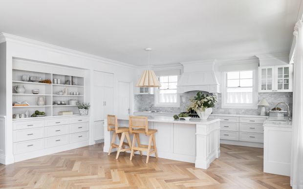 Bright, spacious white kitchen with extensive built-in cabinetry, open display shelving, a central island with timber stools, marble-look benchtops and splashback, and large multi-pane windows. The room has a refined classic coastal feel, with herringbone timber flooring, decorative ceiling mouldings, and a warm neutral palette.