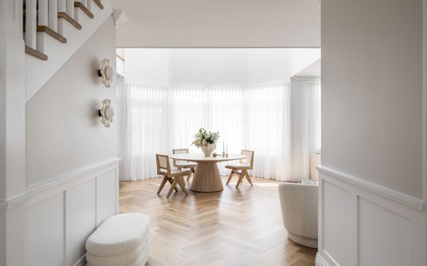 Bright, refined dining area viewed from a white-panelled hallway, with a round timber dining table and sculptural chairs set before floor-to-ceiling sheer curtains. The space features herringbone timber flooring, white wainscoting, a stair with timber treads, and a softly curved upholstered chair in the foreground.