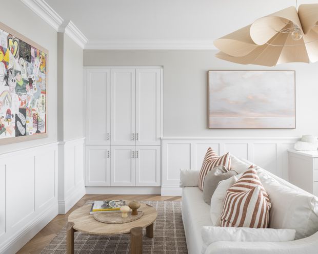 Bright, refined living room with white wall panelling, built-in paneled cabinetry, decorative crown moulding, a light upholstered sofa, striped cushions, a round timber coffee table, layered neutral rugs, and soft contemporary artwork. A sculptural beige pendant light and warm timber flooring add texture and warmth.