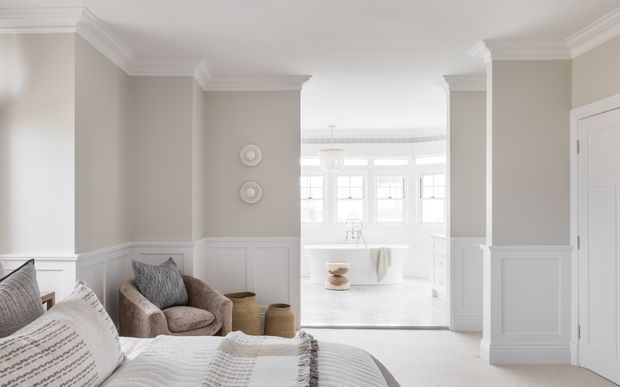 A bright, refined bedroom with a partially visible upholstered bed in the foreground, a comfortable armchair and small side tables in a recessed sitting area, and a freestanding bathtub visible through the open passage beyond. The room features pale neutral walls, white wainscoting, substantial decorative cornices and door trim, and soft natural light.