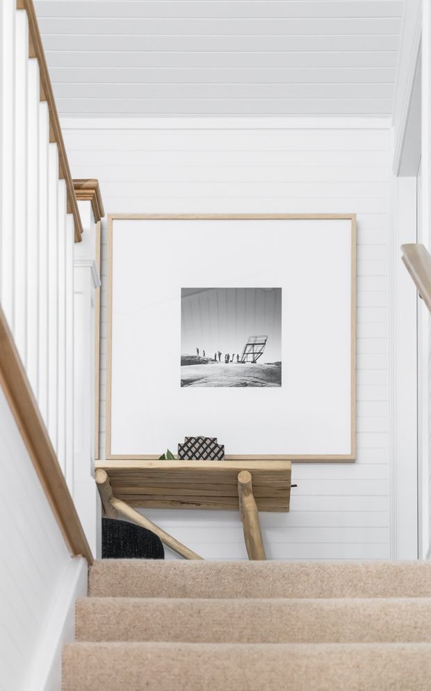 A bright stairway landing with beige carpeted steps, white vertical wall panelling, and a white horizontal-panel ceiling. Timber handrails and a rustic timber console table frame a large monochrome artwork, creating a clean coastal-inspired interior.