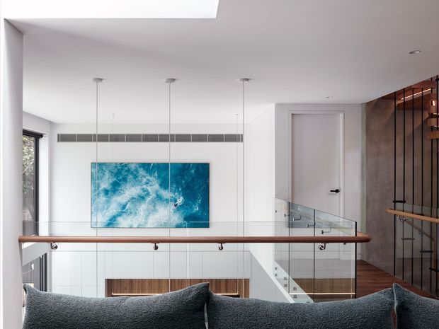View from an upper-level landing or stairway overlooking a double-height contemporary living space. A glass balustrade and timber handrail frame the foreground, with a large blue artwork, linear wall grille, recessed fireplace, white pivot-style door, and a dark timber-and-metal screened feature wall visible. The space has crisp white surfaces, concealed lighting, and clean modern detailing.
