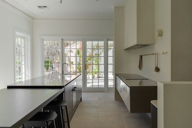 Bright contemporary kitchen with a long dark island and breakfast bar, integrated sink and tap, flat-panel cabinetry, induction cooktop, and a sculptural rangehood. White walls and trim, large steel-framed glazed doors and windows, and pale stone-look flooring create a clean, airy interior with views to a planted courtyard.