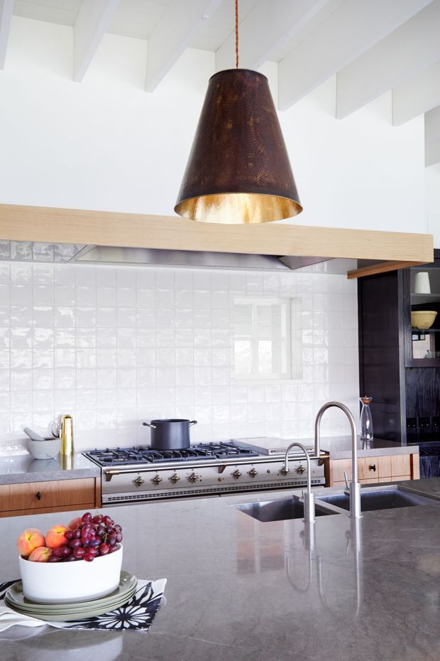 Bright, refined kitchen featuring a large grey stone island with an undermount double sink and brushed metal gooseneck tap, extensive stainless-steel range cooker, white glossy square-tile splashback, timber cabinetry, dark built-in shelving, and a broad light timber overhead beam. White exposed ceiling beams and a large aged copper pendant add character and contrast.