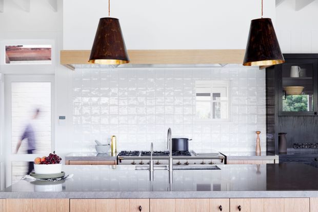 Bright contemporary kitchen with a large central island, white glossy textured tile splashback, stainless-steel range and sink, pale timber cabinetry, and dark timber overhead and side joinery. Two dark pendant lights provide contrast, while a dining or living area is suggested at the left edge.