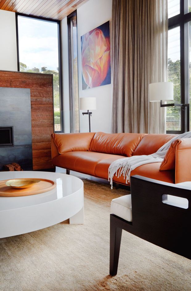 A bright, contemporary living room with floor-to-ceiling black-framed glazing, warm timber ceiling boards, and a timber-and-dark-stone fireplace surround. An orange leather sectional sofa faces a glossy white round coffee table, with a black-framed lounge chair in the foreground. Full-height neutral curtains, wool carpeting, sculptural lamps, and colourful abstract artwork complete the refined interior.