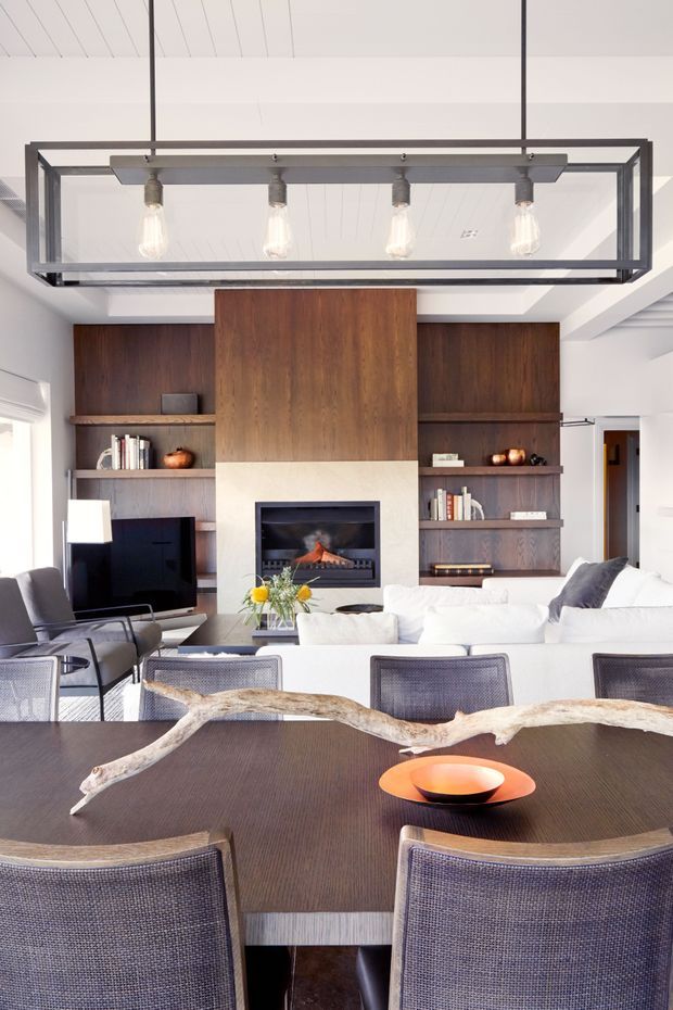 Bright open-plan living and dining room with a contemporary built-in timber feature wall, integrated shelving, and a central fireplace. A large rectangular glass-and-metal pendant hangs over the dining table, while a white sectional sofa, dark upholstered dining chairs, and sculptural driftwood centerpiece create a refined, warm interior.