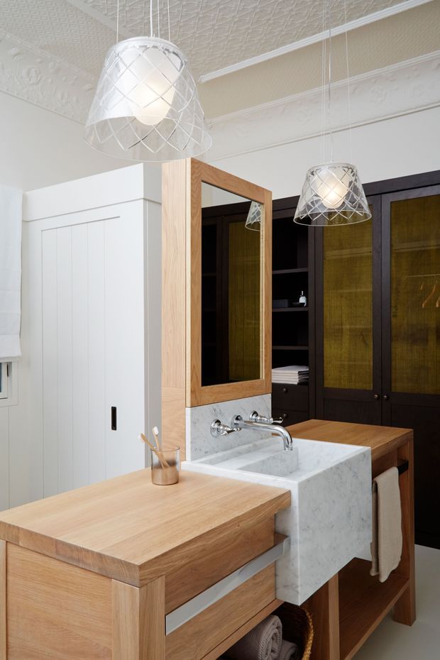 A refined bathroom vanity area featuring a natural timber double-sided vanity with a white marble or stone basin and splashback, chrome wall-mounted tapware, and a timber-framed mirror. Dark timber cabinetry with textured glass panels provides storage in the background. Decorative pendant lights hang from an ornate pressed-metal patterned ceiling, while white paneled walls and detailed cornices contribute a traditional character balanced with contemporary joinery.