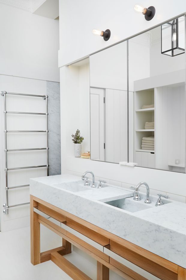 Bright, refined bathroom featuring a large double vanity with a white marble or marble-look benchtop, integrated twin basins, warm timber cabinetry and open base supports. A large wall-to-wall mirror reflects white built-in storage and neatly folded towels. The room also includes a polished chrome heated towel rail, white walls, pale stone wall tiling, and modern black-and-brass wall and pendant lighting.
