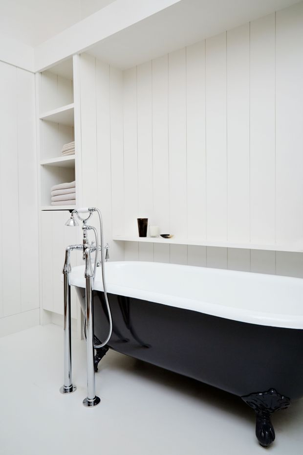 Bright, minimalist bathroom featuring a freestanding clawfoot bathtub with a glossy black exterior and white interior, polished chrome floor-mounted bath taps and hand shower, white vertical wall panelling, integrated open shelving with folded towels, and a white built-in ledge.
