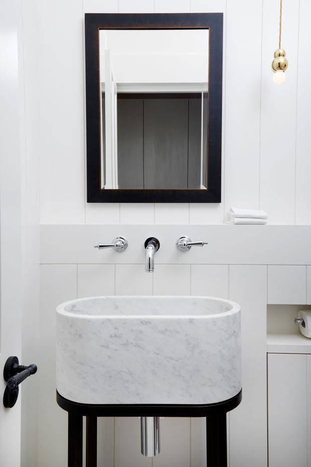 A refined bathroom vanity vignette featuring a freestanding oval white marble basin on a dark timber or stained-wood stand, wall-mounted chrome tapware, a large dark-framed rectangular mirror, and white vertical wall panelling. A small pendant light and built-in open shelving with folded towels are visible.