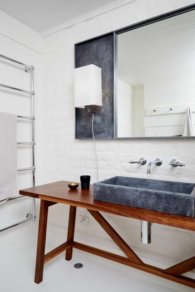 Bright contemporary bathroom featuring a freestanding timber console supporting a deep dark grey stone vessel basin, exposed wall-mounted chrome tapware, white-painted brick walls, a large black-framed mirror, chrome heated towel rail, and white painted ceiling boards. The natural timber base and dark basin create contrast against the predominantly white, high-key interior.