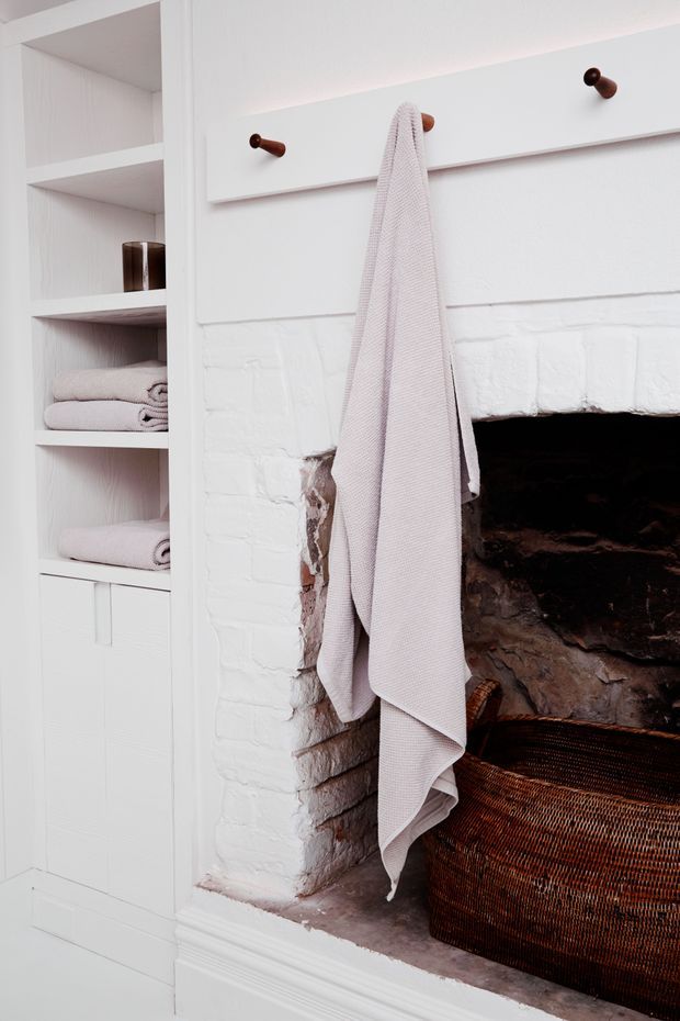 A close-up interior detail showing a white-painted brick fireplace with a dark firebox, a woven brown basket, and a pale towel hanging from a simple wooden peg rail. Built-in white shelving beside the fireplace holds neatly folded towels and a small dark vessel, creating a clean, warm, practical storage feature.