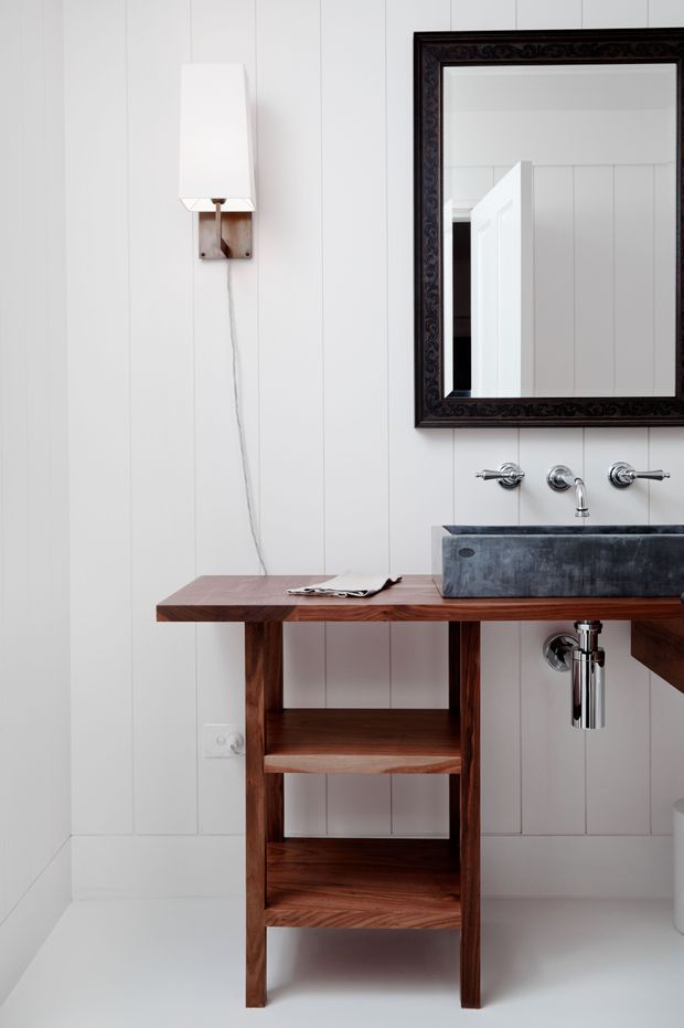 A compact, refined bathroom with white vertical wall panelling and a white floor. A wall-mounted white sconce sits above a floating dark timber vanity with open shelving, supporting a rectangular grey stone basin and polished chrome wall-mounted tapware and trap. A large ornate dark-framed mirror dominates the upper wall.