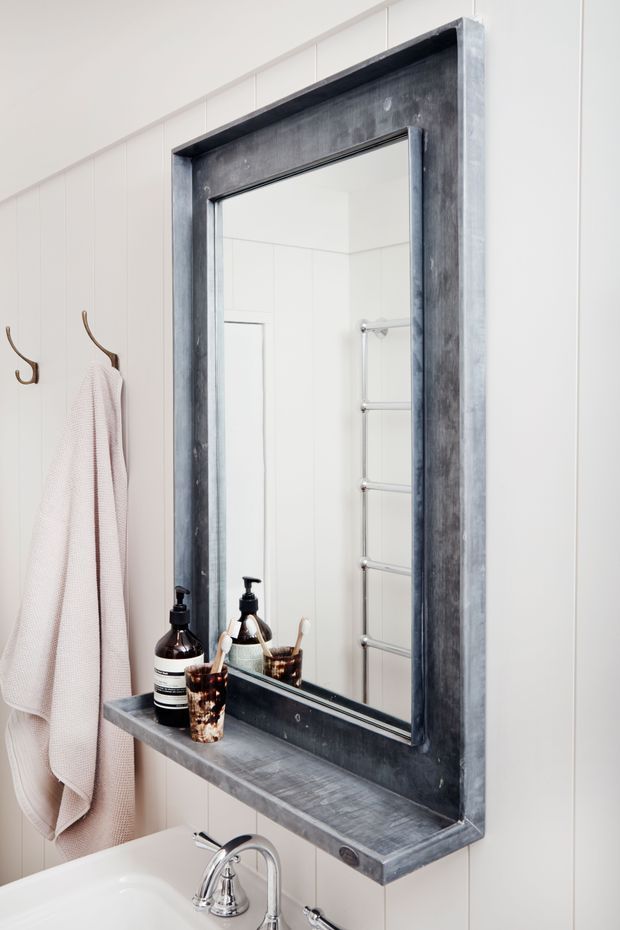 Close-up of a bright bathroom vanity with a white ceramic basin and chrome tap beneath a large rectangular mirror in a substantial dark grey timber frame with an integrated shelf. White vertical wall panelling, bronze wall hooks, a pale pink towel, and dark-toned bathroom accessories create a clean, softly contrasting look.