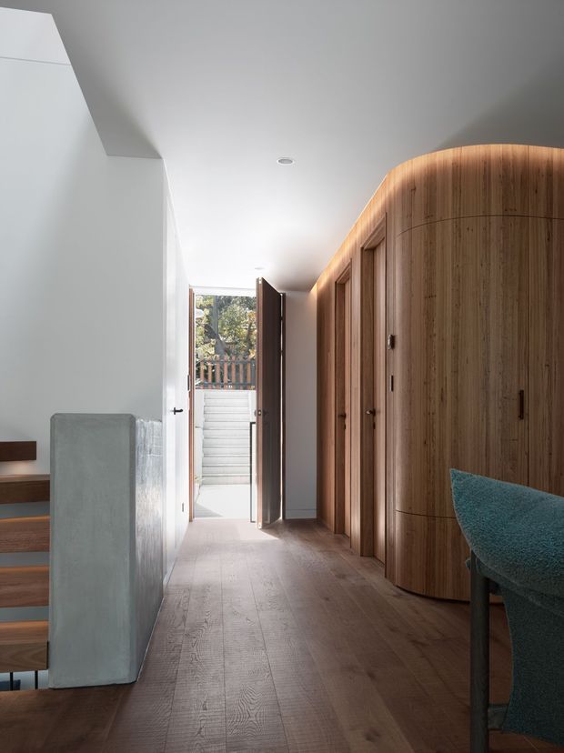 A bright, minimalist residential hallway or entry passage with warm timber flooring and a large curved timber wardrobe or built-in storage wall on the right. White walls and ceiling create a clean, airy setting. A partially visible staircase with timber treads and a pale solid balustrade is on the left, while an open door leads outdoors at the far end. A teal upholstered chair is partially visible in the foreground.