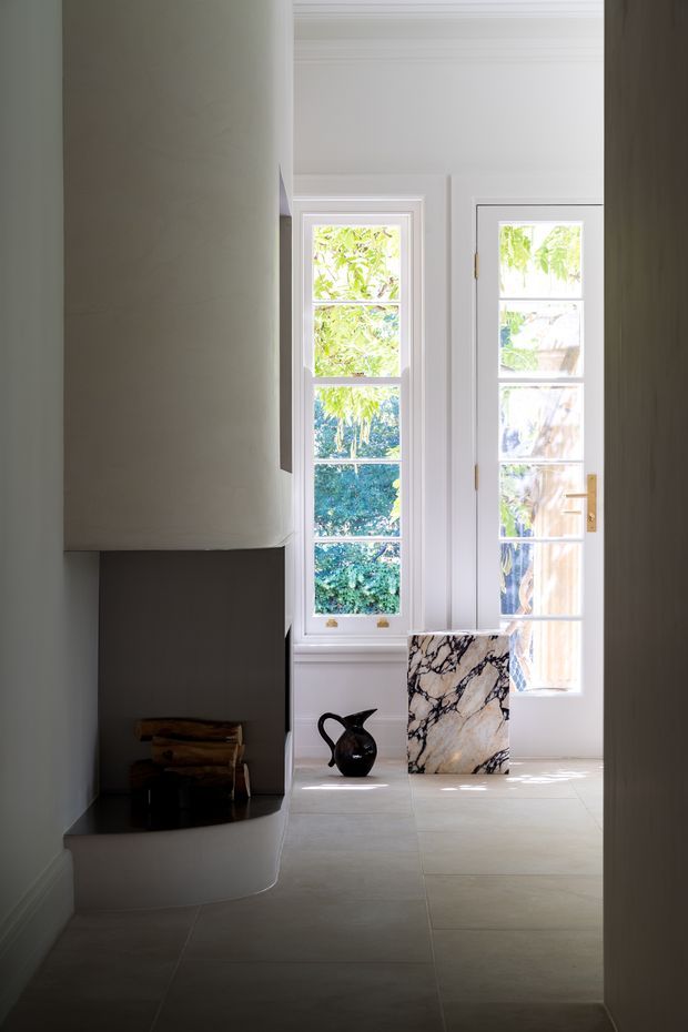 A bright, refined interior passage or living area with a sculptural curved fireplace and dark hearth on the left, tall white-framed multi-pane windows and a glazed door at the rear, pale large-format floor tiles, and a striking veined marble slab leaning against the wall. Minimal decorative objects and abundant garden views create a calm, contemporary atmosphere.