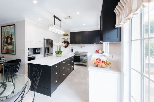 Bright contemporary kitchen with a large white island featuring dark cabinetry, integrated appliances, white perimeter cabinetry, pale tiled flooring, and a broad glazed door or window with layered Roman blinds. A small dining area is visible at the left, along with pendant and recessed lighting and decorative floral accents.