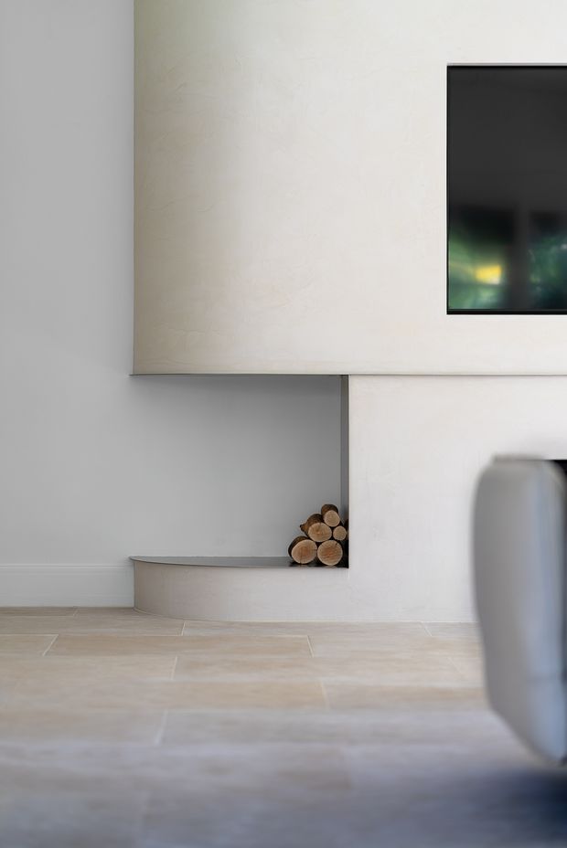 Minimalist interior view featuring a pale textured plaster wall, a wall-mounted black television, and a sculptural recessed display shelf holding stacked firewood logs. The room has large light beige stone or porcelain floor tiles and soft neutral finishes, with part of a rounded upholstered chair or sofa visible in the foreground.