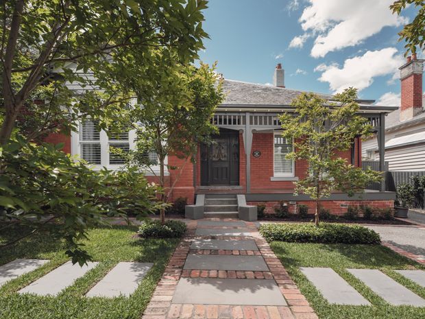 Front exterior of a single-storey red-brick heritage-style house with a symmetrical entry facade, dark timber front door, decorative white verandah screening, slate-like pitched roof, brick chimneys, and a landscaped garden with a brick-and-concrete path. A neighbouring modern structure is visible at the right edge.