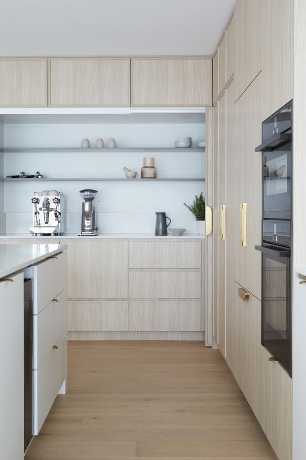 A bright, compact contemporary kitchen with full-height light timber-grain cabinetry, integrated black-glass appliances, brushed gold handles, open shelving, and a pale stone benchtop. A coffee machine and grinder sit along the rear counter, while a peninsula extends into the foreground. The room has light timber flooring, white walls, and a clean, streamlined fit-out.