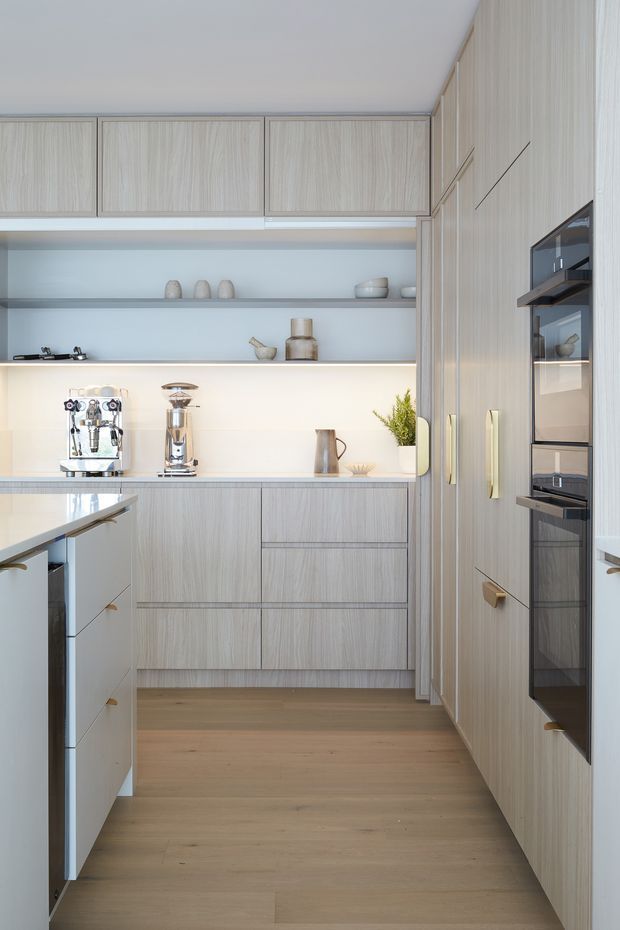 Bright, compact contemporary kitchen with full-height pale timber-grain cabinetry, integrated black-glass ovens, open display shelving, a pale stone or composite benchtop, and warm light timber flooring. A coffee machine and grinder sit on the rear counter, while a partially visible island with drawers occupies the foreground. The restrained palette and concealed storage create a clean, refined look.