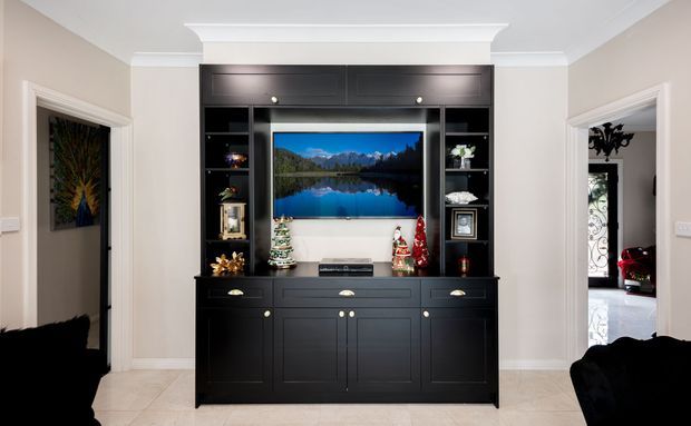 A symmetrical living room media wall with a large black built-in entertainment cabinet, central television, open display shelving, drawers and cupboards. The room has warm off-white walls, white decorative ceiling cornice, pale tiled flooring, and doorways opening to adjacent spaces. Small festive decorations are arranged on the cabinetry.