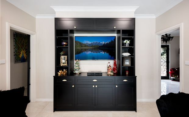 A refined living room media wall featuring a large black built-in entertainment cabinet with upper storage, open display shelving, drawers, cupboards, and a central television. The room has warm neutral painted walls, white decorative crown moulding and ceiling trim, pale tiled flooring, and doorways opening to adjacent spaces. Traditional detailing is paired with a dark, formal built-in joinery unit.