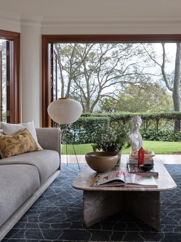 A refined living room with a large grey upholstered sofa, sculptural floor lamp, patterned dark rug, and rounded stone or marble coffee table. Full-height timber-framed glazing opens views to a manicured garden, while decorative objects and cushions add warmth and character. The room has a calm, contemporary-classic atmosphere with strong indoor-outdoor connection.
