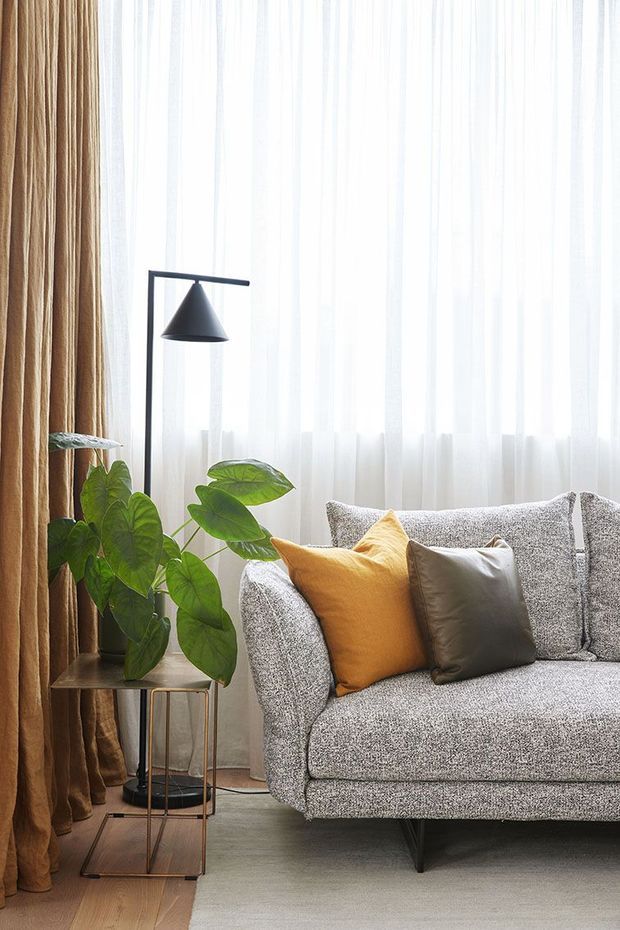 A close-up interior view of a softly styled living room corner, featuring a textured light-grey sofa with mustard and dark taupe cushions, a slim black floor lamp, a small metal-framed side table, and a leafy indoor plant. Full-height sheer white curtains and warm brown drapes frame the bright window, with a neutral rug and timber-toned floor creating a calm, contemporary atmosphere.