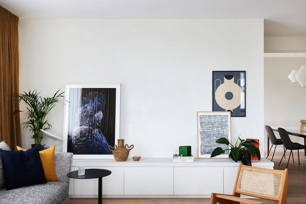 Bright contemporary living room with a low white built-in media cabinet, grey textured sofa, navy and mustard cushions, indoor plants, framed artwork, and a woven cane lounge chair. Timber curtains and warm wood flooring add natural warmth, while a dining area is visible through the opening at right.