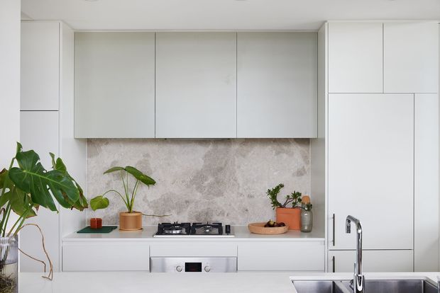 A bright, minimalist contemporary kitchen with streamlined white handleless cabinetry, a pale stone or concrete-look splashback, integrated appliances, a gas cooktop, and a stainless-steel sink and tap. Indoor plants and a few natural decorative elements add warmth to the otherwise restrained palette.