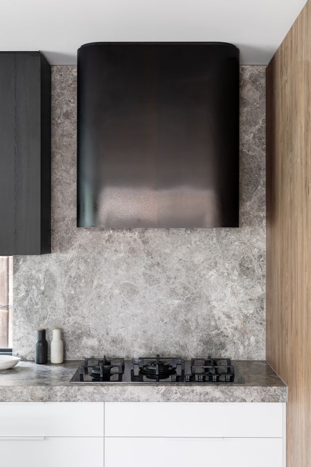 A close-up view of a contemporary kitchen cooking zone, featuring a three-burner black gas cooktop set into a grey stone-look benchtop and matching full-height splashback. A large black curved rangehood dominates the upper wall, flanked by dark cabinetry on the left and warm timber cabinetry on the right. White lower cabinetry provides contrast, with small bottles and a partial sink visible at the edge.