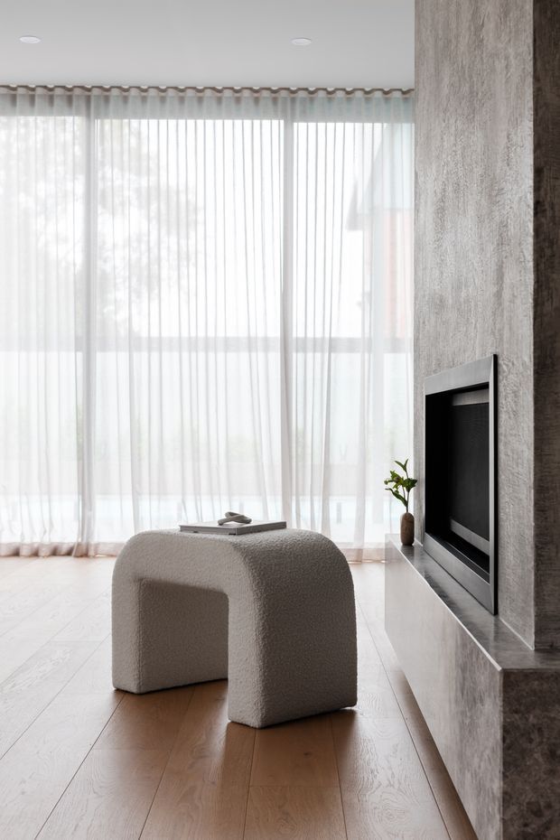 A bright, minimalist living room with floor-to-ceiling sheer curtains, pale timber flooring, and a sculptural boucle ottoman in the foreground. A recessed flat-screen television is integrated into a textured grey stone or concrete feature wall with a low built-in console. The room has abundant soft natural light and restrained neutral finishes.