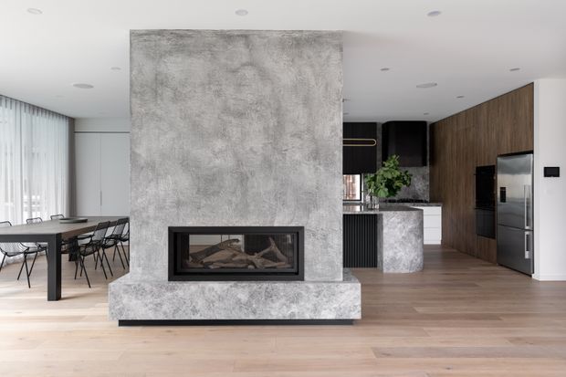 A contemporary open-plan living space centered on a large textured concrete fireplace feature wall with a built-in horizontal firebox. A dining area with a dark table and chairs sits to the left, while a sleek kitchen with dark timber cabinetry, a stone island, integrated appliances, and a stainless-steel refrigerator is visible behind the fireplace. Pale timber flooring, white walls, recessed lighting, and extensive sheer-curtained glazing create a bright, refined interior.