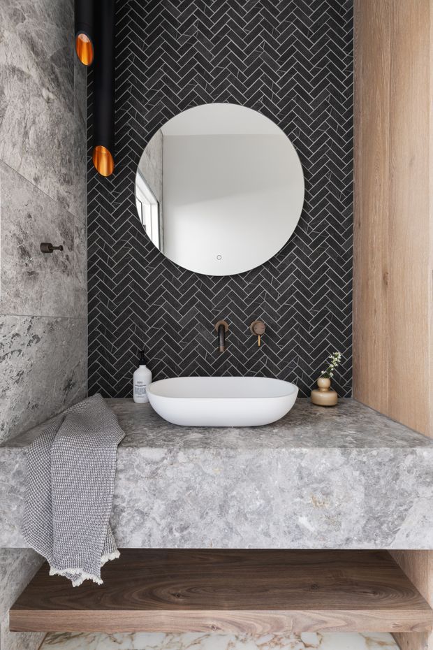 A refined contemporary bathroom vanity featuring a white vessel basin on a grey stone benchtop, a round mirror, dark charcoal herringbone tiled backsplash, grey stone wall surfaces, timber cabinetry and open shelving, and warm copper-toned wall lights.