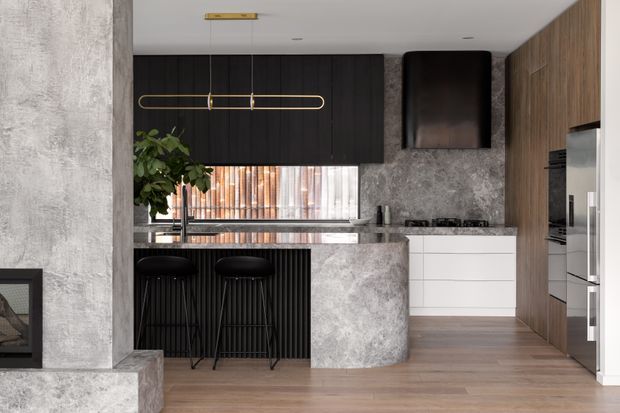 A contemporary kitchen with a dark vertically panelled island base, pale textured stone benchtops and splashback, integrated white drawers, timber-toned cabinetry, and warm timber flooring. A curved island end faces two black bar stools, while a linear brass pendant hangs above. A fireplace edge is visible at left, and a window provides filtered natural light behind the sink.