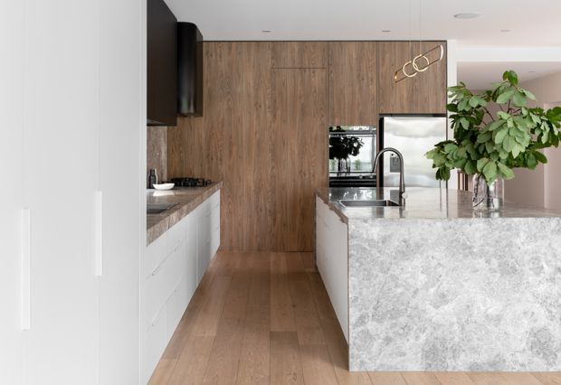 A contemporary galley-style kitchen viewed from the entry, featuring full-height warm timber cabinetry and matching timber flooring, white lower cabinets, dark stone benchtops, and a large waterfall-edge island with a pale stone surface. Stainless steel appliances, an undermount sink with a black tap, integrated lighting, and a leafy indoor plant complete the refined scheme.