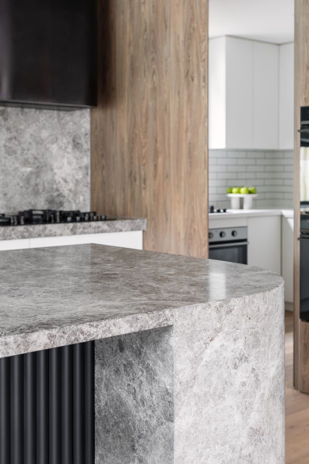 Close-up view of a contemporary kitchen centred on a large curved stone island with a waterfall edge and dark fluted base. The kitchen features a grey veined stone splashback and benchtop, vertical timber-look cabinetry or panelling, white upper cabinetry, a light grey subway-tile splashback, integrated appliances, and black detailing.