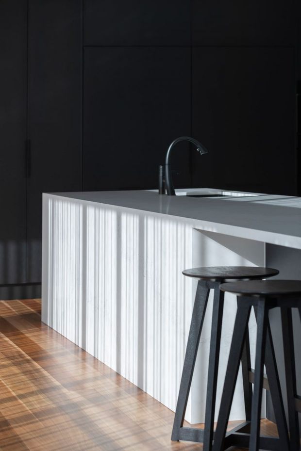A dark, minimalist kitchen featuring a large white island with a vertically ribbed or fluted front, integrated sink and dark curved faucet. Tall matte-black cabinetry forms the backdrop, while black bar stools sit at the island over warm timber flooring. Strong sunlight creates crisp linear shadows across the island and floor.