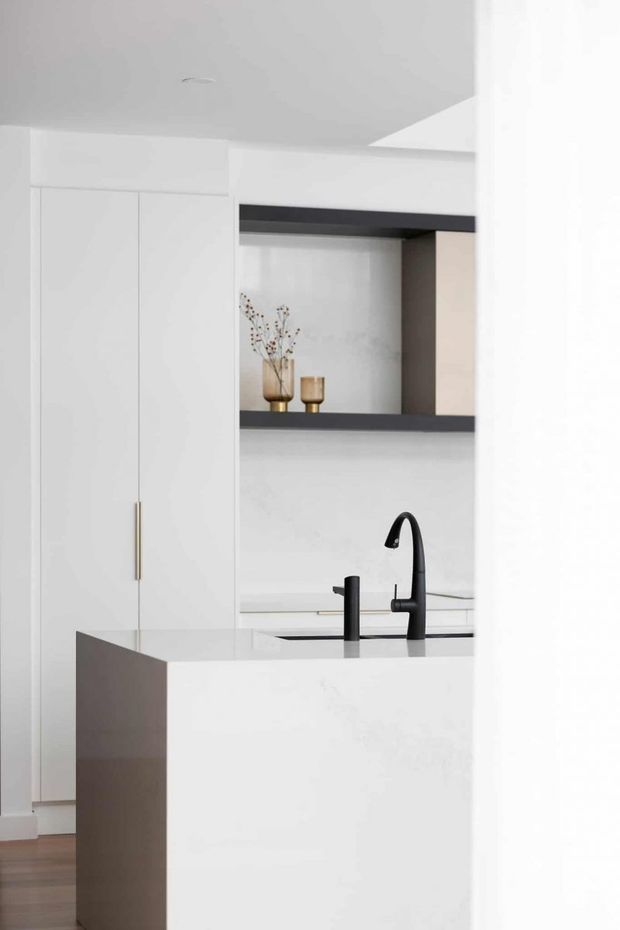 A bright, minimalist kitchen viewed from an adjacent space, featuring tall white handleless cabinetry with a slim brass-toned pull, a white island or peninsula, pale benchtops, and a matte black gooseneck tap with matching secondary fixture. Open dark-framed shelving displays small neutral-toned decorative objects. Warm timber flooring is visible at the edge of the room.