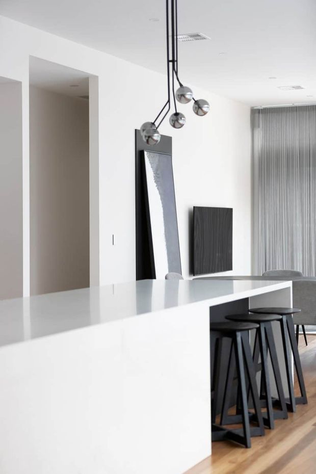 Bright minimalist open-plan kitchen and dining area with a large white island, black bar stools, pale timber flooring, dark wood wall paneling, vertical blinds, and a sculptural cluster pendant light. The space has clean white walls and a restrained black-and-white palette.