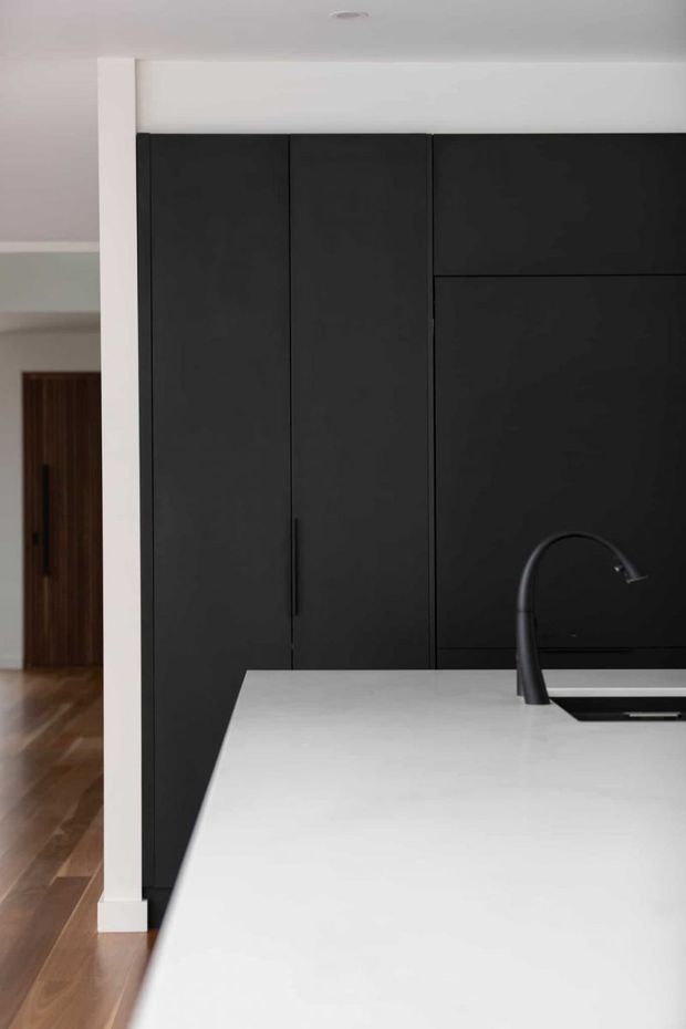 A minimalist contemporary kitchen viewed from the island, featuring full-height matte black cabinetry with integrated handles, a large white island benchtop, and a black gooseneck tap with inset sink. Warm timber flooring and a dark timber door are visible in the adjoining hallway.